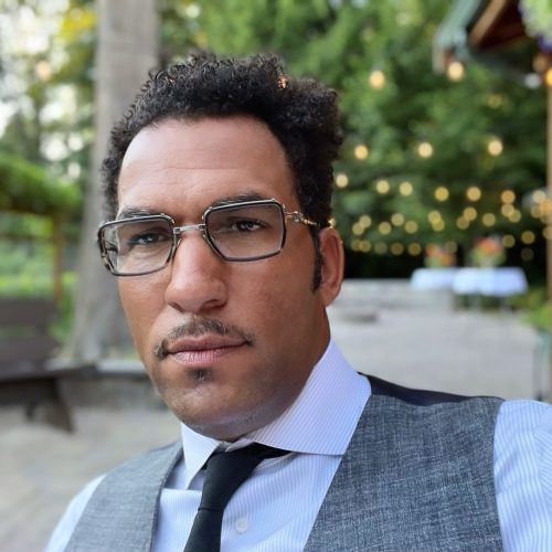 Man wearing glasses and a gray vest, outdoors with greenery in background.